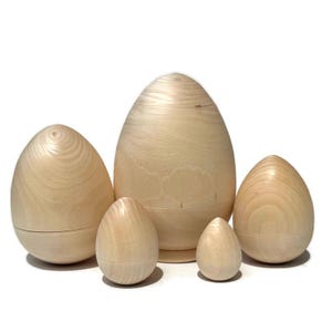 May include: Five unfinished wooden egg-shaped nesting dolls of varying sizes. The eggs are light-colored, with visible wood grain. The largest egg is approximately 7 inches tall, while the smallest is about 2 inches.