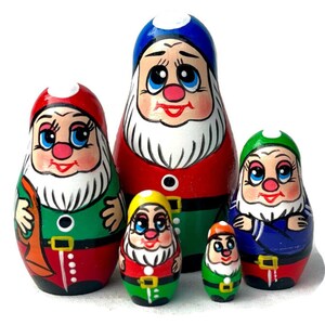 Op de afbeelding: Een set van vijf matroesjka-poppen, elk geschilderd om op een dwerg te lijken. De poppen hebben rode, groene, blauwe en gele hoeden, met witte baarden en lachende gezichten. De grootste pop is ongeveer 13 cm hoog.
