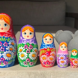May include: A set of seven traditional Russian nesting dolls, or matryoshka dolls. The dolls are painted with colorful floral patterns and have smiling faces. The largest doll is blue with white flowers, the next is green with white flowers, the next is red with white flowers, the next is purple with white flowers, the next is green with white flowers, the next is blue with white flowers, and the smallest doll is red with white flowers.