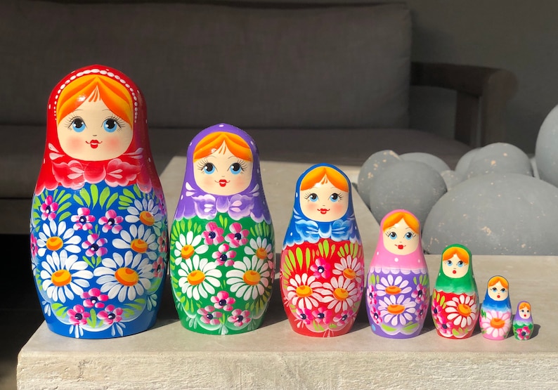 May include: A set of seven traditional Russian nesting dolls, or matryoshka dolls. The dolls are painted with colorful floral patterns and have smiling faces. The largest doll is blue with white flowers, the next is green with white flowers, the next is red with white flowers, the next is purple with white flowers, the next is green with white flowers, the next is blue with white flowers, and the smallest doll is red with white flowers.
