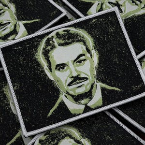 Puede incluir: Parche rectangular con un retrato de un hombre con bigote, en tonos verdes y negros. La imagen tiene un aspecto texturizado y desgastado. El parche está bordeado con costuras blancas. Varios parches son visibles.
