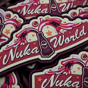 May include: A collection of Nuka World patches. Each patch features a maroon bottle with a white and red splash design, the words "Nuka World" in white, and orange star accents. The patches have a cream-coloured background.