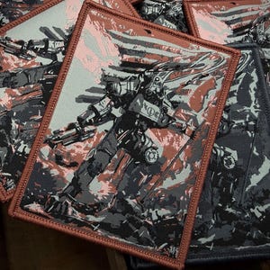 May include: Rectangular patches with a detailed illustration of a futuristic robot in shades of gray, black, and red. The patches have a brown border and a textured appearance, suggesting a woven or embroidered design. Several patches are visible.