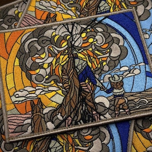 Peut inclure: Écusson brodé avec un motif de vitrail représentant une figure en manteau marron et chemise bleue, un cow-boy et un arbre. L'écusson a une bordure marron et un fond coloré avec des teintes orange, jaune et bleu.
