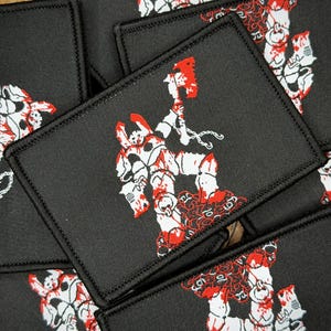 Può includere: Una toppa rettangolare nera ricamata raffigurante una figura in armatura bianca che impugna un'ascia rossa. La figura si erge sopra un mucchio di teschi rossi. Il design presenta dettagli bianchi e rossi con un bordo nero cucito.
