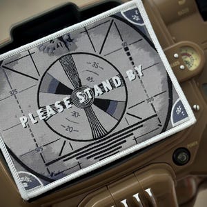 Peut inclure: Un patch rectangulaire avec un motif de mire gris et noir, avec le texte "PLEASE STAND BY". Le patch est placé sur un appareil marron avec des cadrans et des boutons. Le patch a une bordure blanche.