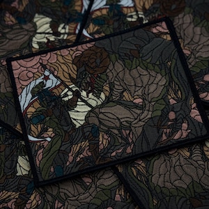 May include: Embroidered patch featuring a detailed illustration of a warrior battling a creature, with a flag in the background. The patch has a stained-glass window effect, with a color palette of greens, browns, and pinks. The patch is square-shaped.