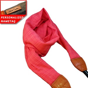 May include: A bright pink camera strap with brown leather ends. The strap is made of a soft, woven fabric. The leather ends have a decorative embossed design. The strap is personalized with a brown leather tag that says "Albert Einstein".