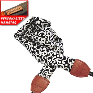 Puede incluir: Correa de cámara negra y blanca con un patrón de paisley. La correa tiene extremos de cuero marrón con un icono de cámara en relieve. La correa se muestra doblada en un bucle.