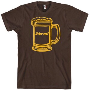 May include: Brown t-shirt featuring a yellow outline of a beer mug with foam and the word "Norm!" in a yellow, retro font. The design is centered on the front of the shirt, offering a casual and humorous style.