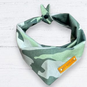 Puede incluir: Un pa&ntilde;uelo para perro de camuflaje en tonos verdes, grises y blancos. El pa&ntilde;uelo est&aacute; atado en la parte superior con un cierre de bot&oacute;n negro. Una peque&ntilde;a etiqueta rectangular de cuero naranja con el texto "RUFF HOPPER CO." est&aacute; adjunta.