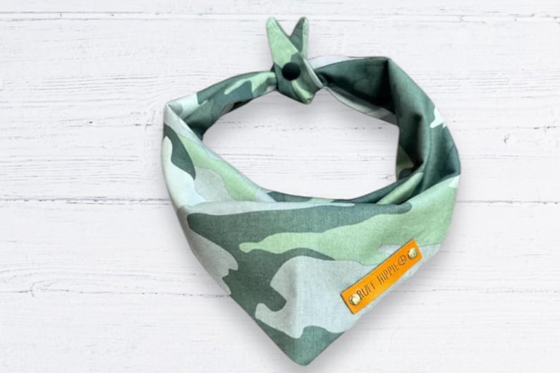 Puede incluir: Un pa&ntilde;uelo para perro de camuflaje en tonos verdes, grises y blancos. El pa&ntilde;uelo est&aacute; atado en la parte superior con un cierre de bot&oacute;n negro. Una peque&ntilde;a etiqueta rectangular de cuero naranja con el texto "RUFF HOPPER CO." est&aacute; adjunta.