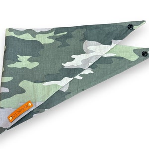 Puede incluir: Un pa&ntilde;uelo para perro con estampado de camuflaje en tonos verdes, grises y blancos. El pa&ntilde;uelo tiene forma triangular con cierres de bot&oacute;n a presi&oacute;n en los extremos. Una peque&ntilde;a etiqueta de cuero naranja con el texto "RUFF HIPPIE CO." est&aacute; adjunta.