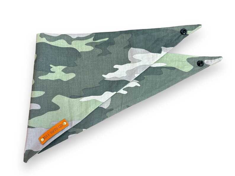 Puede incluir: Un pa&ntilde;uelo para perro con estampado de camuflaje en tonos verdes, grises y blancos. El pa&ntilde;uelo tiene forma triangular con cierres de bot&oacute;n a presi&oacute;n en los extremos. Una peque&ntilde;a etiqueta de cuero naranja con el texto "RUFF HIPPIE CO." est&aacute; adjunta.