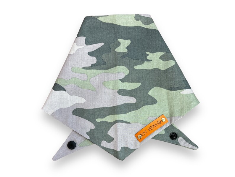 Puede incluir: Bandana para perros con estampado de camuflaje en tonos verdes, grises y blancos. La bandana es triangular y tiene un cierre de bot&oacute;n a presi&oacute;n. Una peque&ntilde;a etiqueta de cuero naranja con el texto "RUFF HIPPIE" est&aacute; adjunta.