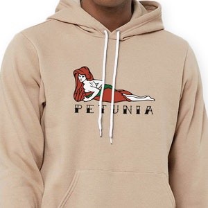May include: A tan hoodie with a graphic of a mermaid with red hair and a green tail. The text "PETUNIA" is printed below the graphic.