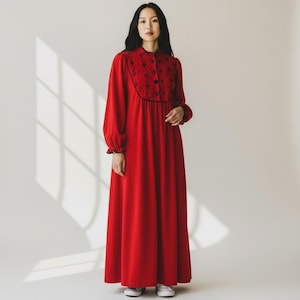 May include: A long, red dress with long sleeves and a high collar. The bodice features a black floral design and black buttons. The dress has a flowing skirt and is worn with white sneakers.