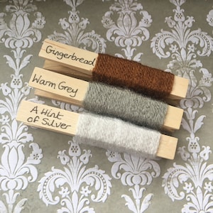 May include: Three skeins of yarn are displayed on wooden pegs. The yarn colours are gingerbread, warm grey, and a hint of silver.