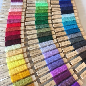 May include: A close-up of a wooden board with yarn samples attached to it. Each yarn sample is labeled with a different city name in the Netherlands. The yarn colors range from bright red to deep purple.