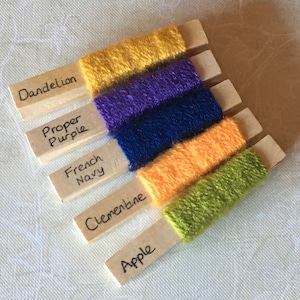 May include: Six wooden spools with yarn wound around them. The spools are labelled with the names of the colours: Dandelion, Proper Purple, French Navy, Clementine, Apple. The yarn colours are yellow, purple, blue, orange, and green.