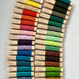 May include: A selection of yarn samples in various colours, each wrapped around a wooden peg. The samples showcase a wide range of shades, including blues, greens, yellows, oranges, and pinks, with handwritten labels indicating each colour.