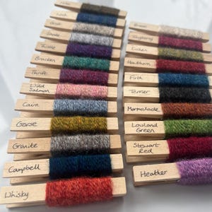 May include: A collection of yarn samples in various colors, including green, red, blue, brown, and gray, are displayed on wooden clothespins. Each clothespin has a handwritten label with a different name, such as "Gorse", "Marmalade", "Lowland Green", and "Stewart Red".