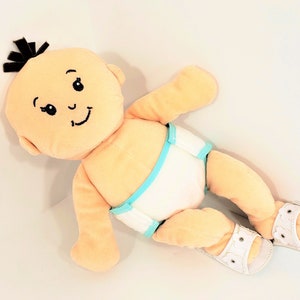 May include: A plush baby doll with peach skin, black hair, and a smiling face. The doll wears a white diaper with a turquoise trim and white sandals with floral accents. The doll is lying on a white surface.