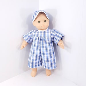 May include: A doll wearing a blue and white plaid romper and matching cap. The doll has a neutral expression and is standing against a white background. The outfit has short sleeves and shorts.