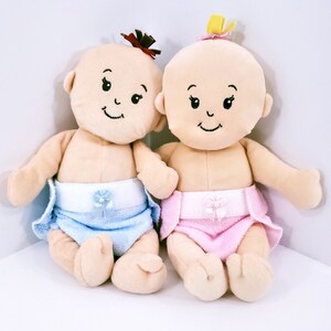May include: Two plush baby dolls, one with a blue diaper and the other with a pink diaper. Both dolls have embroidered facial features and are sitting on a white surface.