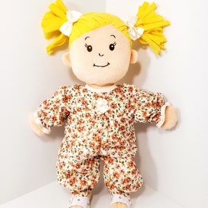 May include: A plush doll with yellow hair styled in pigtails, adorned with white bows. The doll wears a floral print jumpsuit with white sandals. The doll's face features embroidered eyes and a small smile, creating a cheerful appearance.