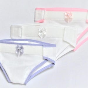May include: Three white cloth diapers with pastel trim and small satin bows. One diaper has pink trim, another has lavender trim, and the third has white trim. The diapers are arranged in a slightly overlapping pattern.