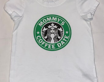 May include: White short-sleeved t-shirt with a green and black graphic. The graphic is a parody of a coffee shop logo, with the text "MOMMY'S COFFEE DATE" encircling a stylized image of a woman. The shirt is made of a soft material.