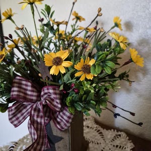 Pu&ograve; includere: Una composizione decorativa di margherite gialle artificiali con centri marroni, fogliame verde e piccole bacche scure. I fiori sono inseriti in un contenitore quadrato in legno con una stella nera intagliata e un fiocco a quadri bordeaux e bianco.