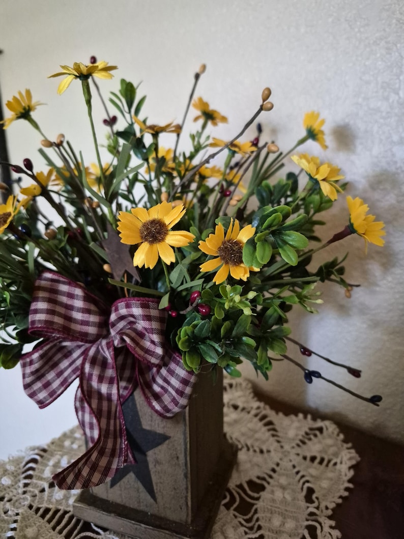 Pu&ograve; includere: Una composizione decorativa di margherite gialle artificiali con centri marroni, fogliame verde e piccole bacche scure. I fiori sono inseriti in un contenitore quadrato in legno con una stella nera intagliata e un fiocco a quadri bordeaux e bianco.