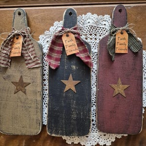 May include: Three rustic wooden cutting boards in shades of tan, black, and burgundy. Each board features a star and a tag with the words "Faith Family Friends." The boards are adorned with fabric bows.