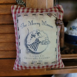 May include: A red and white checkered fabric pillow with a vintage-style illustration of a mouse holding a plate of cookies. The text on the pillow reads "The Merry Mouse Cookie Co. Delivering Goodies Since 1810".
