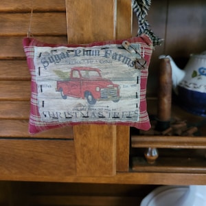May include: A red and white plaid pillow with a vintage-style print of a red pickup truck carrying a Christmas tree. The print reads "Sugar Plum Farms, Spruce Fir Cedar, Est. 1898, One Mile Down Old Mill Rd, Christmas Trees".