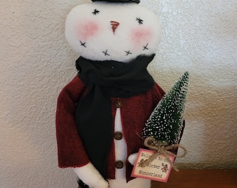 May include: A handmade snowman doll wearing a red and black coat, a black scarf, and a black top hat. The snowman is holding a small green Christmas tree and a sign that says "Welcome to our Winter Wonderland".