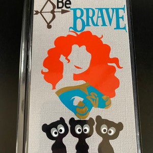 May include: A gray notebook with a black cover and a silver background. The cover features a silhouette of a red-haired woman with a blue dress and a gold crown. The text "Be Brave" is written in blue letters above the woman. Three black bears with large eyes are at the bottom of the cover.