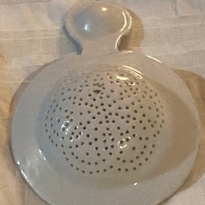 May include: White ceramic strainer with a handle and a round, perforated base. The strainer is designed for rinsing small items.