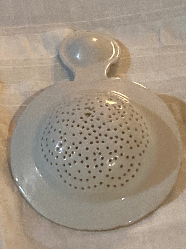 May include: White ceramic strainer with a handle and a round, perforated base. The strainer is designed for rinsing small items.