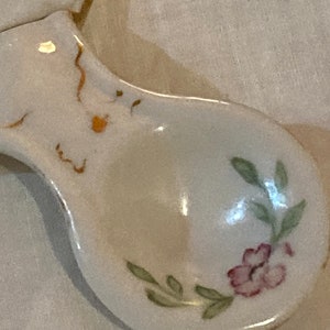 May include: A white ceramic spoon with a floral design. The spoon has a pink flower with green leaves and a gold trim.