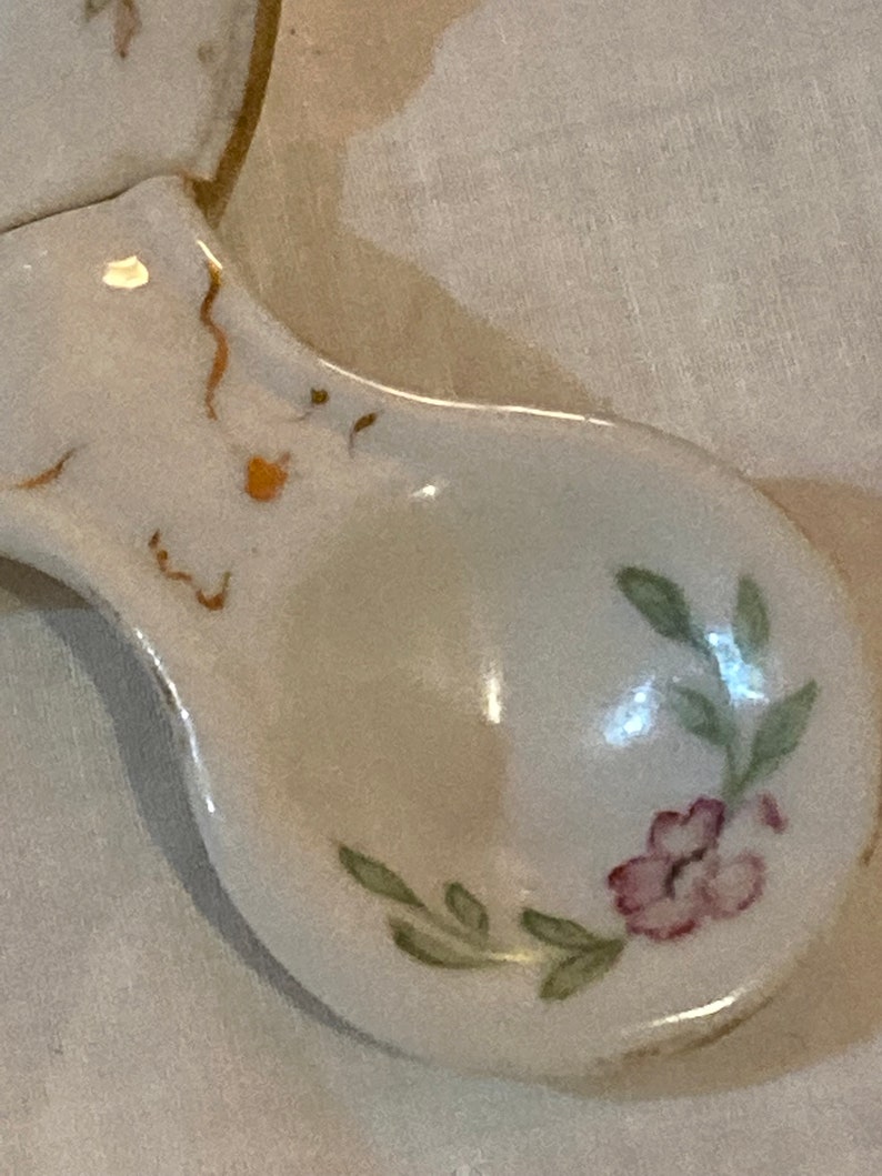 May include: A white ceramic spoon with a floral design. The spoon has a pink flower with green leaves and a gold trim.