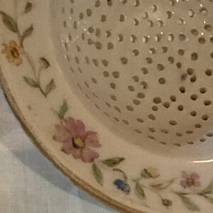 May include: A white ceramic colander with a floral design. The colander has a round shape and a handle. The floral design is in shades of pink, blue, and green.