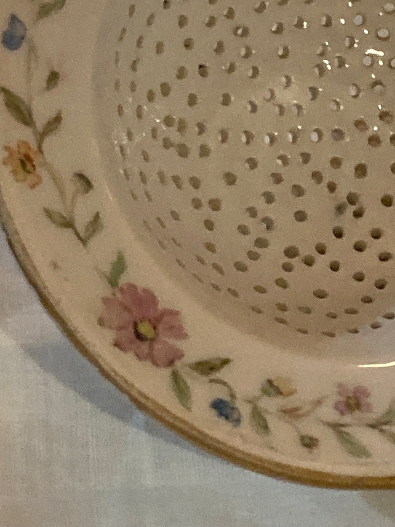 May include: A white ceramic colander with a floral design. The colander has a round shape and a handle. The floral design is in shades of pink, blue, and green.