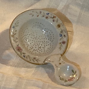 May include: A white ceramic tea strainer with a floral design and a handle. The strainer has a round, perforated bowl and a small, round handle.
