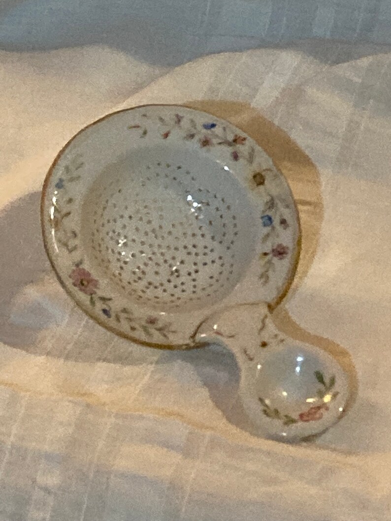 May include: A white ceramic tea strainer with a floral design and a handle. The strainer has a round, perforated bowl and a small, round handle.