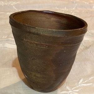 Könnte beinhalten: Eine braune Keramiktasse mit einem breiten Rand und einem leicht ausgestellten Boden. Die Tasse hat ein rustikales, handgemachtes Aussehen.