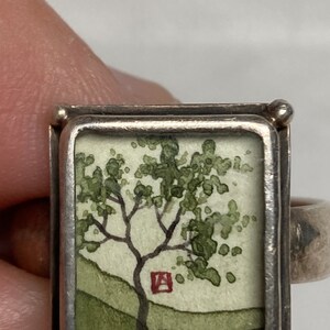 May include: A silver ring with a square frame containing a watercolor painting of a green tree with a red square in the background.