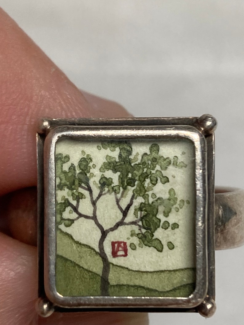 May include: A silver ring with a square frame containing a watercolor painting of a green tree with a red square in the background.